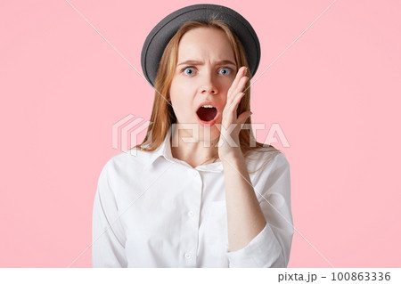 Close up portrait of shocked female with stunned expression, keeps mouth wide opened, hears shocking news from interlocutor, wears elegant shirt and black stylish hat, isolated on pink background Close up portrait of shocked female with stunned expression, keeps mouth wide opened, hears shocking news from interlocutor, wears elegant shirt and black stylish hat, isolated on pink background 100863336