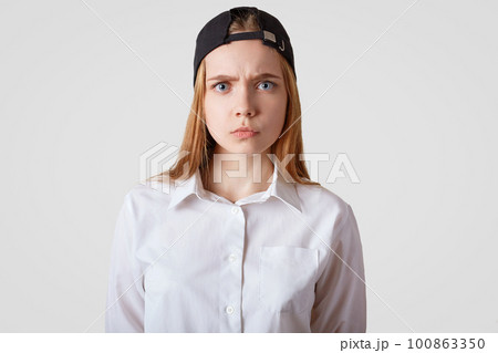 Waist up portrait of grumpy female model with sullen expression, wears black cap and elegant shirt, being dissatisfied with something, isolated over white studio background. People and emotions 100863350
