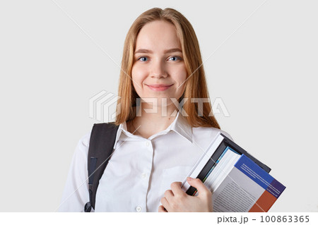Sweet adorable blond haired young female poses isolated over white background, holding many books in both hands, looking directly at camera, looks pleased and confident, having black bagback. 100863365