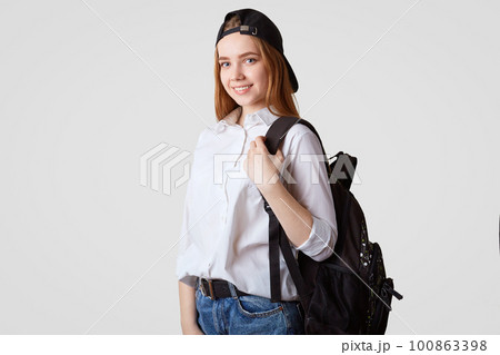 Happy schoolgirl in black cap and white shirt, carries rucksack with books, ready for starting new studying year, poses against white background. College student returns back home from classes Happy schoolgirl in black cap and white shirt, carries rucksack with books, ready for starting new studying year, poses against white background. College student returns back home from classes 100863398