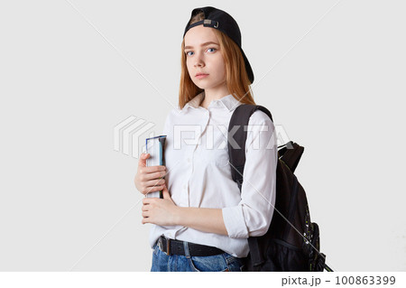 Horizontal shot of student girl standing isolated over white background, holding paper folders in hands, wearing blouse, black cap and backpack, looking at camera. Education and teenager concept. 100863399