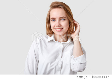 Photo of pleasant looking woman with toothy smile, blue eyes, healthy skin, has nice hairstyle, dressed in elegant formal shirt, ready for meeting, isolated over white background, has natural beauty 100863437