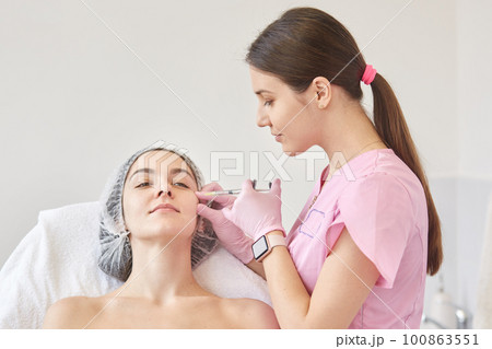 Photo of young oman getting collage injection in her face at clinic. Cosmetologist wears pink medical gown filling female wrinkles by hyaluronic acid. Woman sits on couh calmly. Anti aging concept. Photo of young oman getting collage injection in her face at clinic. Cosmetologist wears pink medical gown filling female wrinkles by hyaluronic acid. Woman sits on couh calmly. Anti aging concept. 100863551
