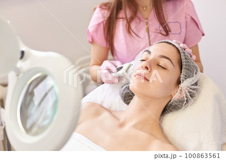 Beautiful woman receiving ultrasound facial peeling. Skin cleansing procedure at beauty spa salon. Cosmetologist makes cleansing treatment for her young client via moder equipment. Skin care concept. Beautiful woman receiving ultrasound facial peeling. Skin cleansing procedure at beauty spa salon. Cosmetologist makes cleansing treatment for her young client via moder equipment. Skin care concept. 100863561