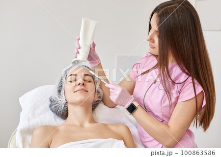 Doctor cosmetologist doing face lifting massage for young brunette woman, client lying on couh woth closed eyes, wearing white towel and medical cap. Aethsetic medicine, cosmetology and beauty concept Doctor cosmetologist doing face lifting massage for young brunette woman, client lying on couh woth closed eyes, wearing white towel and medical cap. Aethsetic medicine, cosmetology and beauty concept 100863586