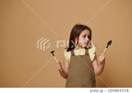 Beautiful 5-6 years old child, adorable little kid girl in khaki dress, holding a garden shovel and rake in her hands, isolated over beige background. Gardening. People, nature and Earth Day concept. Beautiful 5-6 years old child, adorable little kid girl in khaki dress, holding a garden shovel and rake in her hands, isolated over beige background. Gardening. People, nature and Earth Day concept. 100864358