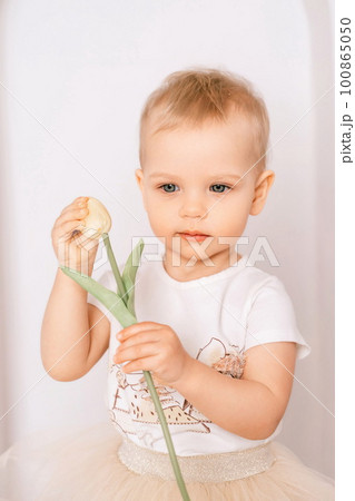 Baby girl elegant dress. A one-year-old girl in a puffy skirt and a white T-shirt poses against the backdrop of a bright room with a yellow tulip in her hand 100865050