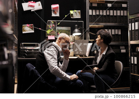 Fbi coworkers working overtime at fraud case, analzing company financial report on tablet computer in arhive room. Senior agent discussing investigation evidence with private detective Fbi coworkers working overtime at fraud case, analzing company financial report on tablet computer in arhive room. Senior agent discussing investigation evidence with private detective 100866215