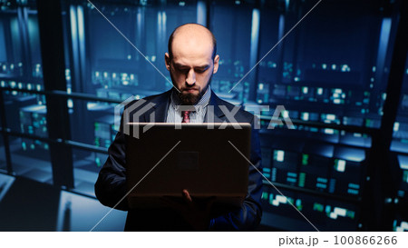 System engineer running hosting server diagnostics with laptop, standing in modern big data center space. IT technician working on professional mainframe hardware. Tripod shot. 100866266