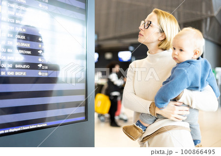 Mother traveling with child, holding his infant baby boy at airport terminal, checking flight schedule, waiting to board a plane. Travel with kids concept 100866495