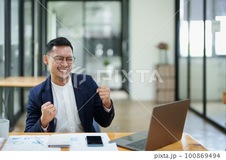 Portrait of an Asian male business owner standing with a computer showing happiness after a successful investment. 100869494