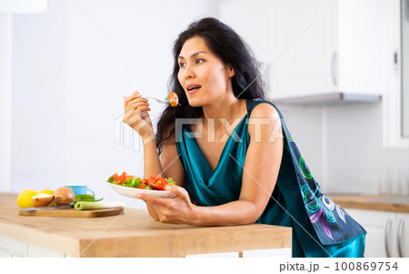 Woman leaning on kitchen table and eating salad 100869754