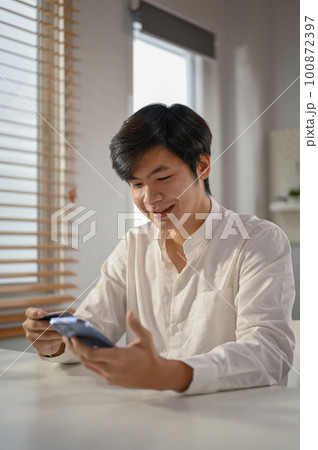 Smiling millennial Asian man using mobile banking application to pay online bills. Smiling millennial Asian man using mobile banking application to pay online bills. 100872397