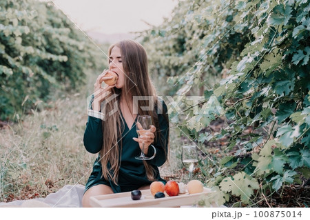 Woman picnic vineyard. Happy woman with a glass of wine at a picnic in the vineyard, wine tasting at sunset and open nature in the summer. Romantic dinner, fruit and wine. 100875014