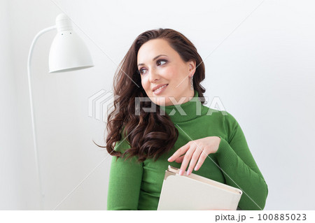 Brunette woman holds book while sitting on chair in white apartment - leisure and free time concept Brunette woman holds book while sitting on chair in white apartment - leisure and free time concept 100885023