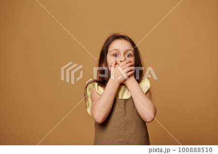 A frightened little girl stands on a beige background, covering her mouth with her hands, feeling fear, afraid to reveal a secret. Stress. Children's rights. Childhood fears. Child protection concept 100888510