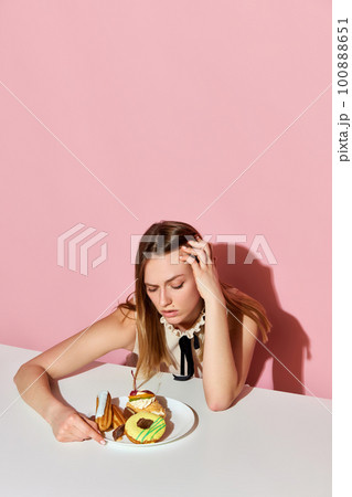 Pretty woman sitting at table with junk food and looking at plate with upset eyes. Concept of diets, food addiction, beauty Pretty woman sitting at table with junk food and looking at plate with upset eyes. Concept of diets, food addiction, beauty 100888651