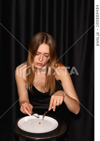 One young weeping girl sitting at table with empty plate and looking away over black background 100889484