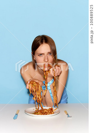 Portrait of adorable young girl eating spaghetti by hands with sad facial expression over blue background Portrait of adorable young girl eating spaghetti by hands with sad facial expression over blue background 100889525
