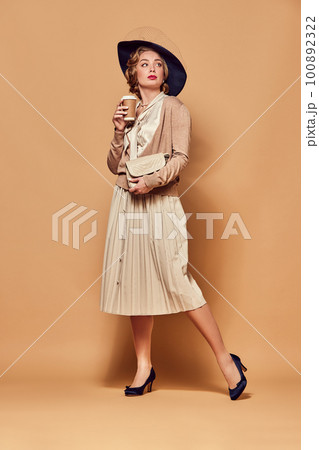 Beautiful woman wearing retro clothes holding cup of coffee and looking away over beige background 100892322