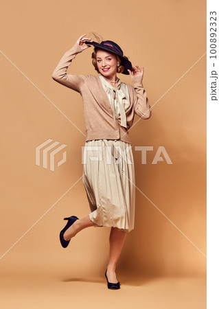 Portrait of charming lady wearing hat smiling at camera over beige background. Music, dance, charm, grace concept 100892323