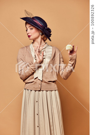 One beautiful woman wearing retro style clothes holding cake with unpleasant facial expression over beige background One beautiful woman wearing retro style clothes holding cake with unpleasant facial expression over beige background 100892326