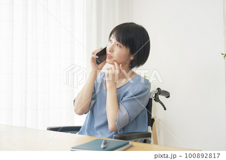 リビングでスマートフォンで電話しながら考える車椅子に乗った女性 100892817