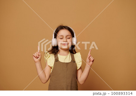 Lovely baby girl enjoys new sountrack, points her index fingers up and smiles, posing with her eyes closed on beige background with copy ad space. People. Children. Entertainment. Modern technologies Lovely baby girl enjoys new sountrack, points her index fingers up and smiles, posing with her eyes closed on beige background with copy ad space. People. Children. Entertainment. Modern technologies 100893314
