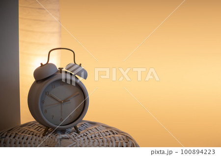 On the wicker lid of the basket stands an analog alarm clock against a background of a lit bedside lamp. On the wicker lid of the basket stands an analog alarm clock against a background of a lit bedside lamp. 100894223