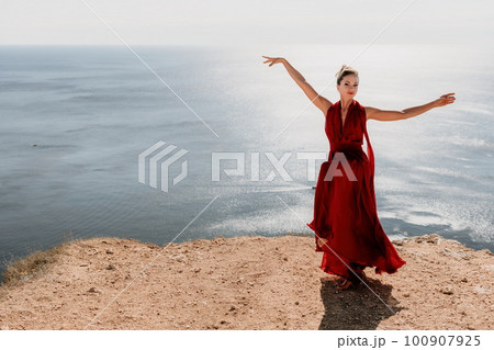 Woman in red dress on sea. Side view a Young beautiful sensual woman in a red long dress posing on a rock high above the sea on sunset. Girl on the nature on blue sky background. Fashion photo. 100907925