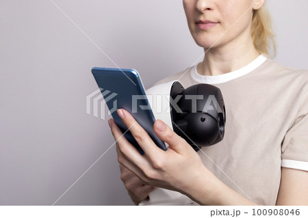 White Female Person Holds IP Wifi Outdoor Security Camera And Looks at Smartphone On White Grey Background. Smart Home, House Surveillance, Protection. Horizontal, closeup 100908046