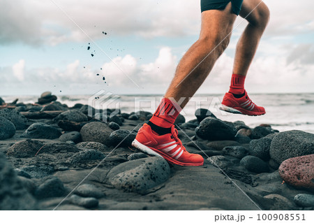 Running shoes of athlete man jogging on beach. Difficult terrain with wet rocks for training outdoors. Runner exercising 100908551