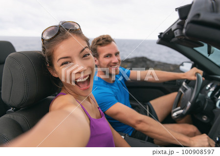 Selfie car travel couple having fun on summer road trip vacation driving to holidays. Asian woman and man excited taking self-portrait photo with mobile phone 100908597