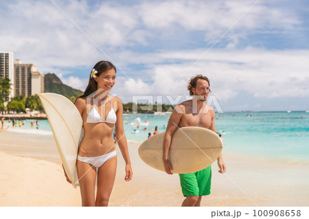 Hawaii Waikiki beach couple tourists surfers happy going surfing. Interracial couple, Asian bikini model woman, Caucasian man walking with surfboards after surf lesson 100908658