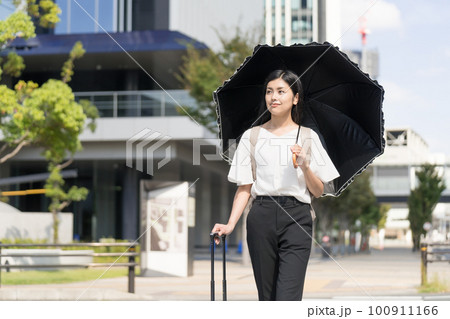 キャリーバックを持って日傘をさしてビジネス街を歩くビジネスウーマン キャリーバックを持って日傘をさしてビジネス街を歩くビジネスウーマン 100911166