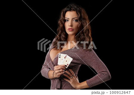 Brunette girl wearing shiny dress posing holding two playing cards in her hand standing against black studio background. Casino, poker. Close-up. 100912647