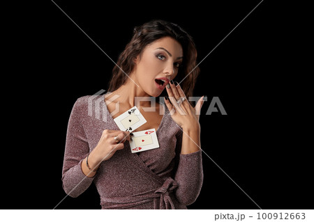 Brunette girl wearing shiny dress posing holding two playing cards in her hand standing against black studio background. Casino, poker. Close-up. Brunette girl wearing shiny dress posing holding two playing cards in her hand standing against black studio background. Casino, poker. Close-up. 100912663