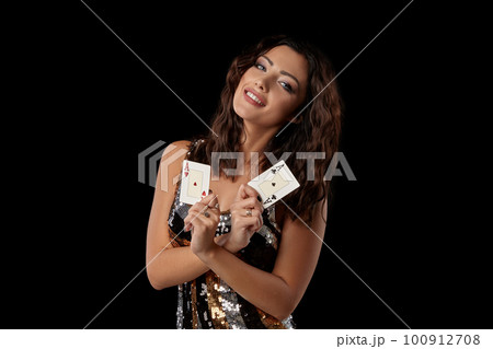 Brunette girl in a colored sequin dress posing holding two playing cards, standing against black studio background. Casino, poker. Close-up. 100912708