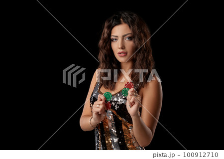 Brunette girl in a colored sequin dress posing holding some chips, standing against black studio background. Casino, poker. Close-up. 100912710