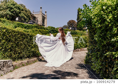Brunette runs white dress park. A beautiful woman with long brown hair and a long white dress runs along the path along the beautiful bushes in the park, rear view Brunette runs white dress park. A beautiful woman with long brown hair and a long white dress runs along the path along the beautiful bushes in the park, rear view 100914279