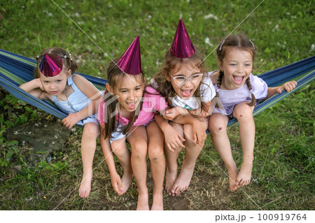Four funny girlfriends in festive pink hats are sitting on a hammock and laughing merrily. Fun and a birthday party for the child. A holiday in the courtyard in nature Four funny girlfriends in festive pink hats are sitting on a hammock and laughing merrily. Fun and a birthday party for the child. A holiday in the courtyard in nature 100919764