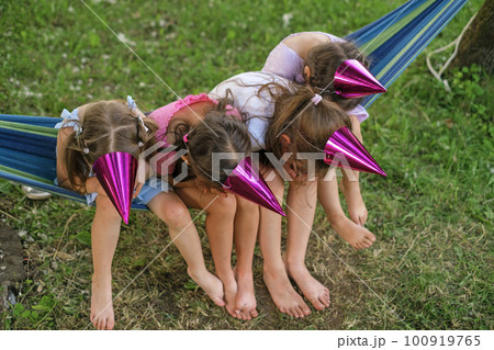 Four humorous girls in festive bright shiny purple hats are sitting on a hammock hiding their faces joking. Fun and birthday party. A holiday in the courtyard in nature on a summer day Four humorous girls in festive bright shiny purple hats are sitting on a hammock hiding their faces joking. Fun and birthday party. A holiday in the courtyard in nature on a summer day 100919765