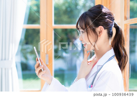 窓際でスマホを使う若い女性医師 窓際でスマホを使う若い女性医師 100936439