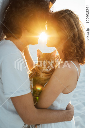 A handsome man with curly hair gently hugs his beloved. They have a cat in their hands A handsome man with curly hair gently hugs his beloved. They have a cat in their hands 100938264