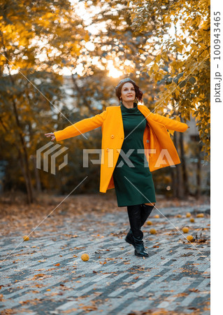 Beautiful woman walks outdoors in autumn. She is wearing a yellow coat and a green dress. Young woman enjoying the autumn weather. Autumn content. 100943465