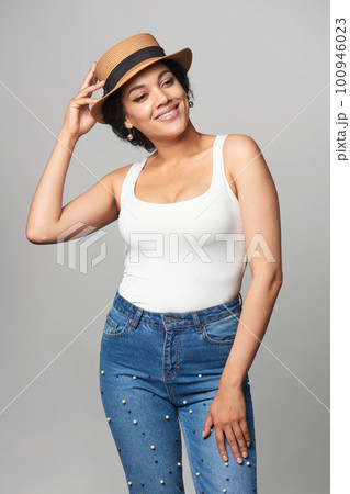 Relaxed casual mixed race woman in jeans and white tank top wearing canotier straw hat 100946023