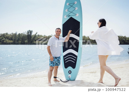 Surfers couple on the beach. Man stand with paddleboard and smiling. He waiting his partner. Young beautiful woman in sunglasses and white srirt running towards her boyfriend. Surfers couple on the beach. Man stand with paddleboard and smiling. He waiting his partner. Young beautiful woman in sunglasses and white srirt running towards her boyfriend. 100947522