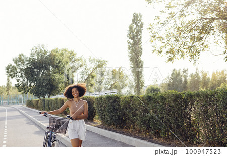 Dark skinned female teenage is driving her bicycle. Young and positive afro american girl in casual cloth smiles sweetly and has a fun. Dark skinned female teenage is driving her bicycle. Young and positive afro american girl in casual cloth smiles sweetly and has a fun. 100947523