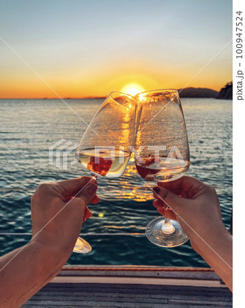 Female hands holding glasses of champagne on board the boat. Sunset sky and sea on the background. Making a celebratory toast with sparkling wine. Female hands holding glasses of champagne on board the boat. Sunset sky and sea on the background. Making a celebratory toast with sparkling wine. 100947524