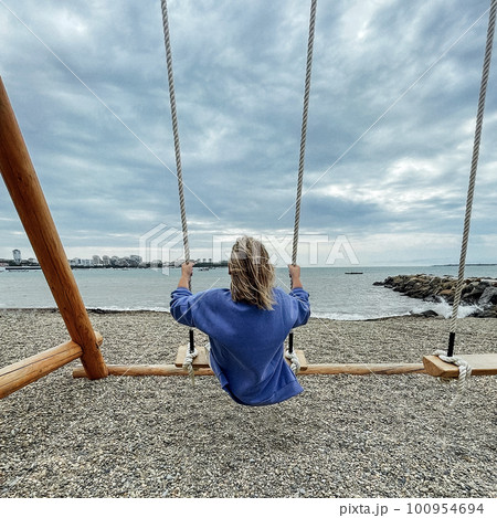 Young blonde girl rides on a wooden swing and sees the sea. View from the back. 100954694
